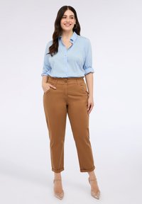 Camicia azzurra chiara con maniche arrotolate, abbinata a pantaloni marroni a sigaretta. L'outfit presenta un tessuto morbido e un design a vita alta.