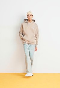Bež dukserica s uvlakom i prednjim džepom, uparena s svijetloplavim trapericama i bijelim tenisicama. Neutralna pletena kapa. Čvrste boje kroz cijeli outfit.