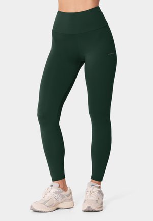 Nogi u ciemnozielonych legginsach z wysokim stanem i jasnobeżowych butach New Balance, stojące na jednolitym jasnym tle.