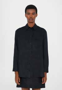 Black long-sleeve shirt with a button front, collar, and single chest pocket featuring embroidered detail. Made of textured fabric.