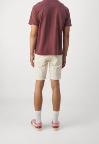 Muž oblečený v vínovém tričku, béžových kraťasech s ohrnutými nohavicemi, bílých ponožkách a červených teniskách Nike, stojí zády k fotoaparátu na jednobarevném pozadí.