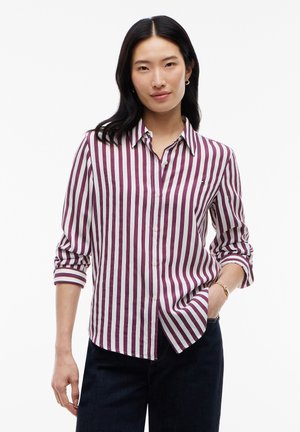 Femme avec de longs cheveux noirs portant une chemise à rayures verticales blanches et bordeaux boutonnée et un pantalon foncé, debout avec une main dans la poche.