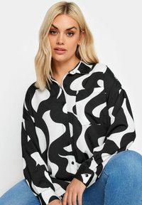Camicia a bottoni bianca e nera con un motivo ondulato, maniche lunghe e una tasca sul petto; indossata con jeans in denim blu.