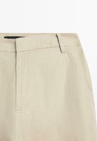 Pantalon beige taille haute avec passants de ceinture et poche avant, gros plan sur la texture du tissu et les détails des coutures.