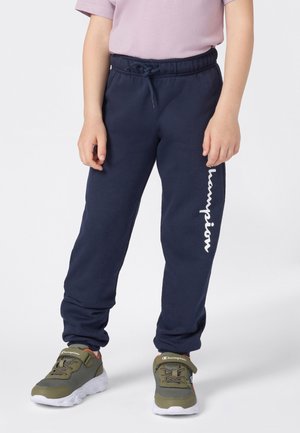 Kind in marineblauwe Champion trainingsbroek en olijfgroene Champion sneakers, staande voor een effen witte achtergrond.