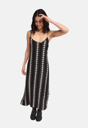 Jeune femme portant une robe noire sans manches avec des motifs floraux roses verticaux et des bottines noires, souriante et touchant ses cheveux.