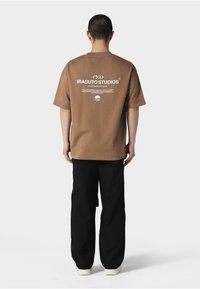 Brun oversized t-skjorte med trykk på ryggen som har teksten "IRASUTO STUDIOS" i hvitt. Kombinert med svarte vide bukser og lyse sko.