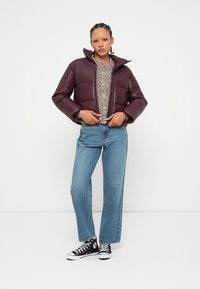 Doudoune bordeaux avec un col montant, associée à un pull beige texturé et un jean large bleu clair. Des baskets noires achèvent le look.