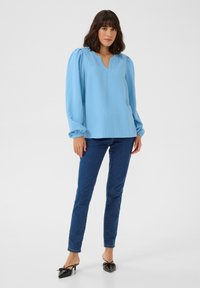 Blouse bleu clair à manches longues avec un décolleté en V, en tissu texturé, et avec des poignets froncés, assortie à un jean ajusté bleu foncé et à des mules à talons noirs.