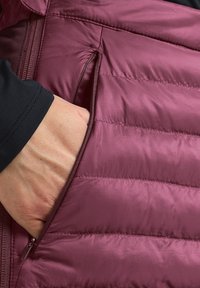 Bordeauxkleurige geïsoleerde jas met horizontale gewatteerde vulling en een ritszak. Close-up van een hand in de zak. Zachte textuur.