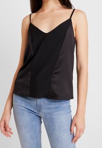 Musta satiininen kimalletoppi, jossa on V-pääntie ja ohuet hihnat. Malli on hieman löysä, ja edessä on vinonauhaompeleita.