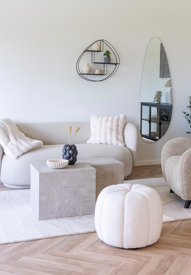 Gekrümmtes beiges Sofa mit strukturierten Kissen, grauem Marmor- couchtisch, weißem Pouf, schwarzem Metallschrank mit Dekoration und einem ovalen Spiegel an einer hellen Wand.