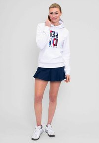 Sudadera blanca con un logotipo gráfico colorido, combinada con una falda plisada azul marino. Zapatillas deportivas blancas completan el conjunto. Textura de tela suave.