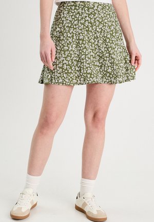 Grøn floral nederdel med små hvide blomster, lavet af letvægtsstof, med en svajet design og en glat tekstur. Kombineret med hvide sneakers.