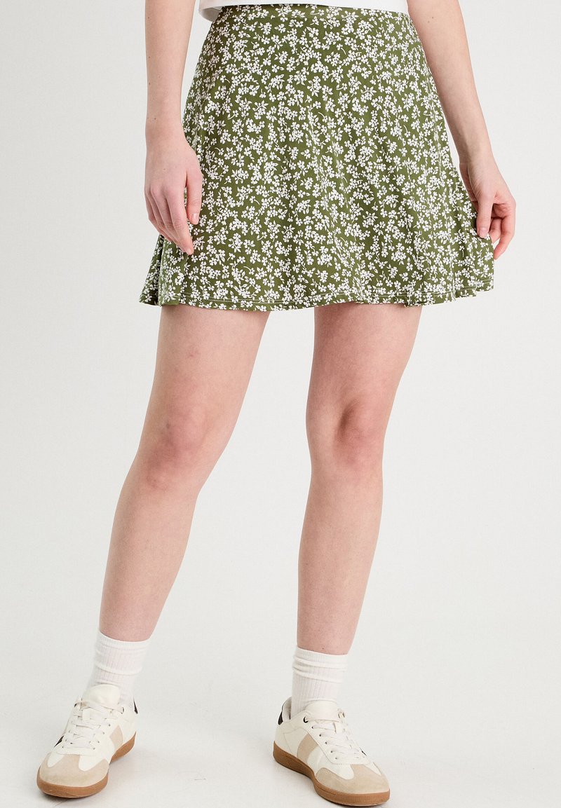 Jupe florale verte avec de petites fleurs blanches, en tissu léger, dotée d'un design évasé et d'une texture lisse. Associée à des baskets blanches.