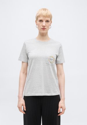 Person med kort blondt hår iført grå t-shirt med blomsterlomme-design og sorte bukser med smalle striber, stående mod en ensfarvet baggrund.