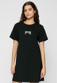 Robe t-shirt en coton noir avec des manches courtes, arborant le texte blanc "pray" en police gothique sur le devant. Design à col rond, coupe décontractée.