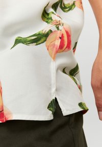 Blusa floral de tejido blanco con ilustraciones de melocotón, hojas verdes, aberturas laterales y detalles de costura en contraste.