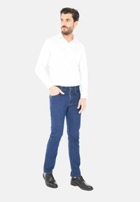 Uomo con capelli scuri che indossa una camicia bianca, jeans blu e scarpe nere, in piedi con una mano in tasca, che guarda di lato.