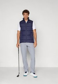 Marinblå vadderad väst med vertikal quiltad design, dragkedja fram, bärs över en vit t-shirt, kombinerad med ljusgrå byxor och vita golfskor.