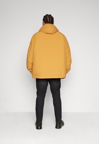 Ett vadderat gult jacka med huva, vida ärmar och en något kortad längd, parat med mörka jeans och svarta skor.