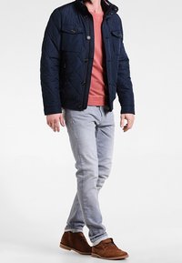 Marinblå quiltad jacka med fyra framfickor, röd skjorta under, ljusblå jeans och bruna mockaskor.