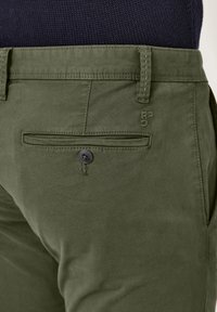 Olivegrüne, leichte Baumwollhose mit einer Gesäßtasche, die einen schwarzen Knopf und ein gesticktes Markendetail in der Nähe des Bundes aufweist.