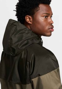 Kurtka odporna na wodę w ciemnooliwkowej zieleni i czerni. Posiada kaptur, gładką teksturę oraz kontrastowe bloki kolorystyczne na ramionach.
