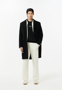 Abrigo largo negro sobre un sudadera negra con logo bordado, pantalones anchos color crema y zapatos blancos con cordones sobre un fondo neutro.