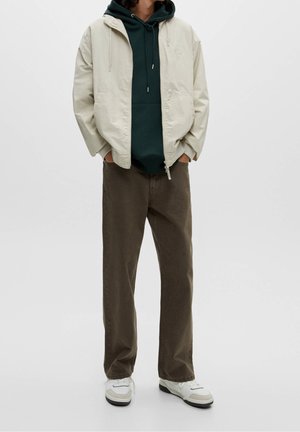 Giacca beige chiaro con zip sopra una felpa verde scuro con cappuccio, abbinata a pantaloni marroni a taglio ampio e sneakers bianche, in piedi contro uno sfondo neutro.