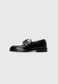 Mocasín de cuero negro con un acento de cuero blanco en la parte delantera, que presenta un detalle de hebilla cuadrada y un tacón bajo apilado. Textura suave.