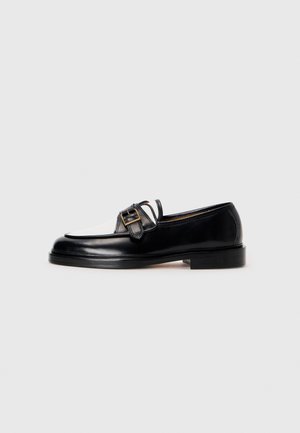 Black leather loafer with a white leather accent at the front, featuring a square buckle detail and a low stacked heel. Smooth texture.