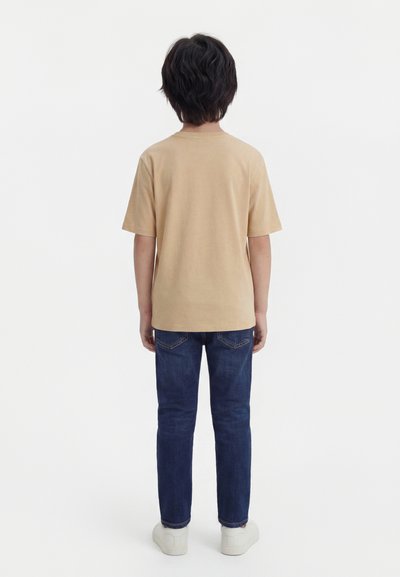 Παιδί με σκούρα μαλλιά, ντυμένο με μπεζ πουκάμισο κοντομάνικο, μπλε τζιν και άσπρα αθλητικά παπούτσια, στέκεται με την πλάτη προς τα εμπρός σε λευκό φόντο.