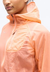 Γυναίκα που φοράει ανοιχτό πορτοκαλί μπουφάν με κουκούλα, φερμουάρ μπροστά και φερμουάρ σε τσέπη στο στήθος, εμφανίζεται από τον λαιμό έως το μέσο του κορμού.