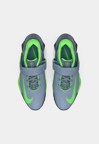 Ténis atléticos cinzentos com um destaque verde brilhante, parte superior em malha texturada, colarinho acolchoado e tira ajustável, demonstrando um design elegante.