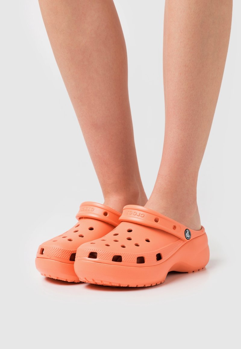 Personne portant des sabots Crocs orange avec trous de ventilation et bride arrière, debout sur un fond clair et uni.