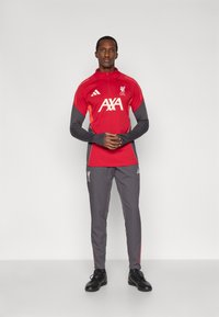 Top de entrenamiento rojo del Liverpool FC con acentos grises, mangas largas y collar con cremallera; combinado con pantalones grises con franjas rojas laterales. Calzado negro.