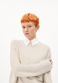 Hellbeiger Pullover mit langen Ärmeln, strukturiertem Strick, getragen über einem weißen Hemd mit Kragen. Kurze, leuchtend orangefarbene Haare. Neutrale Hintergrund.