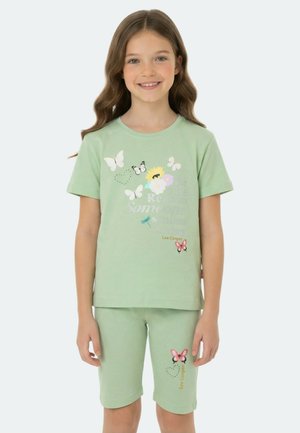 Jeune fille portant un short et un t-shirt assortis vert clair ornés de papillons, de fleurs et du texte « Lee Cooper », souriant à la caméra.