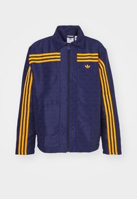 Mörkblå jacka med präglat mönster, som har tre gula ränder på varje ärm och en liten gyllene logotyp på vänster bröst.