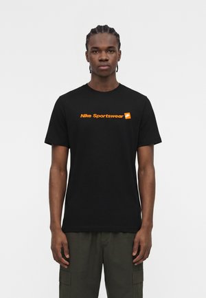 Jeune homme aux cheveux tressés portant un t-shirt noir Nike Sportswear avec un logo orange et un pantalon cargo sombre sur un fond uni.
