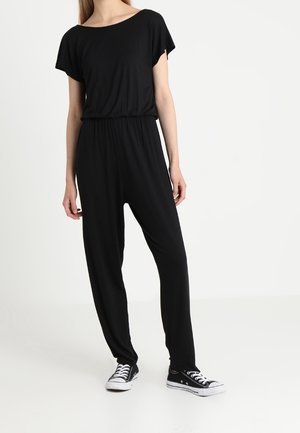 Personne portant une combinaison noire ample à manches courtes et des baskets basses noires avec semelles et lacets blancs.