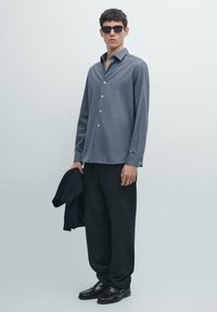 Šedá košile na knoflíky s límečkem, v kombinaci s tmavými kalhotami s širokými nohavicemi. Černé sluneční brýle, černé boty a v ruce drží tmavou bundu.