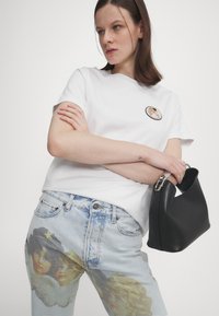 Donna che indossa una t-shirt bianca con una patch di angelo e jeans azzurri chiari decorati con volti di cherubini, mentre tiene una borsa nera su uno sfondo bianco.