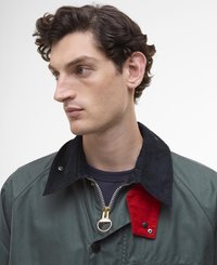 Casaco verde com fecho à frente, colarinho de veludo cotelê preto em contraste, um patch vermelho em destaque e detalhes em dourado. Textura suave, design utilitário.