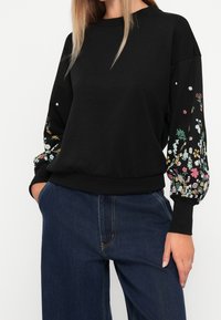 Femme portant un sweat-shirt noir à manches longues avec des motifs floraux colorés sur les manches et un jean en denim bleu foncé, les mains dans les poches.