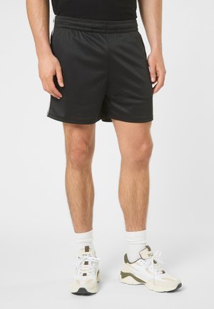 Mand iført sorte atletikshorts, hvide crew-sokker og hvide FILA-sneakers, der står foran en ensfarvet baggrund.