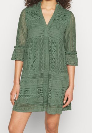 Kobieta ubrana w zieloną sukienkę koronkową do kolan z rękawami do łokcia, dekoltem w serek oraz misternymi kwiatowymi i geometrycznymi wzorami.