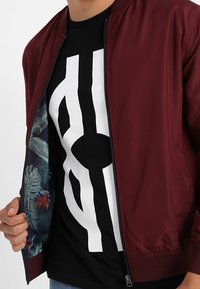 Chaqueta bomber burdeos con cierre de cremallera, que muestra un forro estampado. Debajo, una camiseta negra presenta un diseño gráfico blanco.