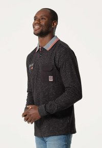 Polo à manches longues noire avec un col rayé contrasté, patte de boutonnage, poche poitrine et détail en tissu texturé.